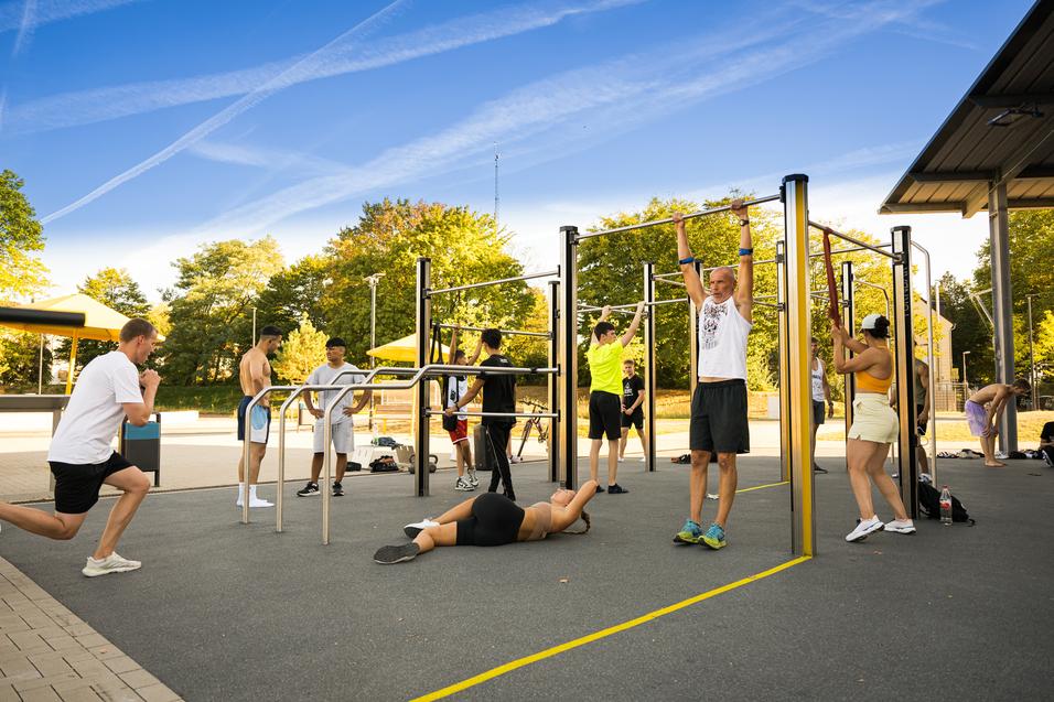 Gruppe von Personen trainiert an Freiluftfitnessgeräten in einem Park unter blauem Himmel.